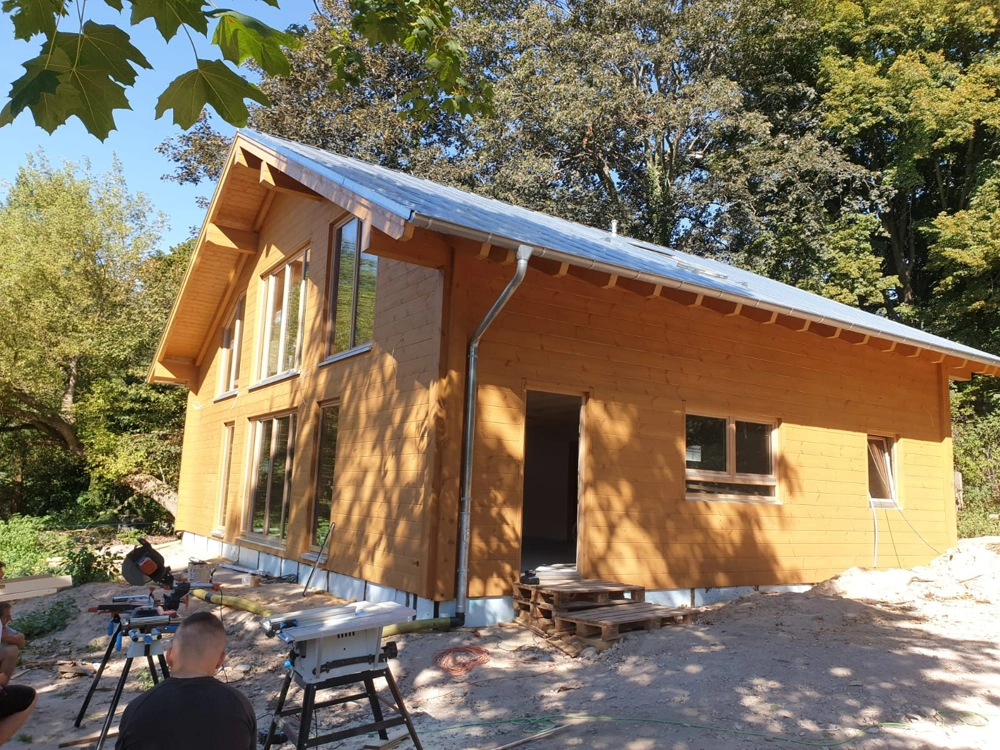 Montage eines Holzhauses mit Bauunternehmen, Tischkreissäge und Handwerkern im Vordergrund