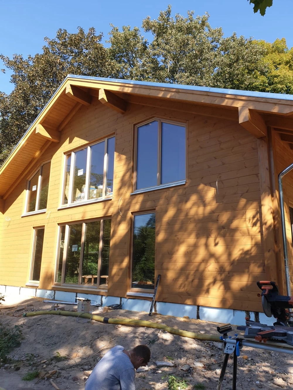 Holzhaus-Rohbau mit großen Fensterfronten, Fundamentarbeiten im Vordergrund und Arbeiter mit Werkzeug – Bauunternehmen