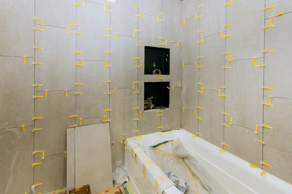Großformatige graue Wandfliesen werden im Badezimmer um eine Badewanne mit gelben Distanzhaltern verlegt – Fliesenleger