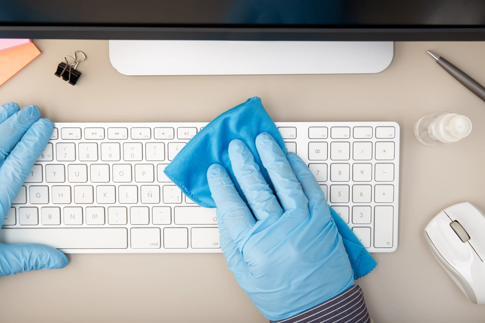Büroreinigung: Mitarbeiter mit blauen Handschuhen reinigt weiße Computertastatur mit Mikrofaser-Tuch