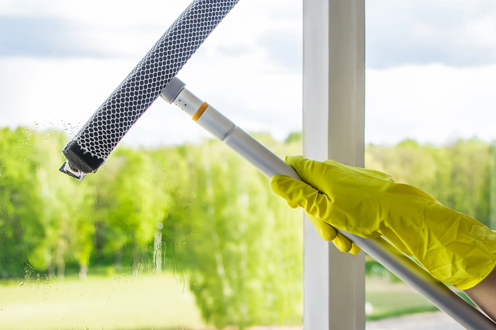 Fensterreinigung: Hand mit gelbem Gummihandschuh entfernt Wasser mit Fensterabzieher vor grünem Hintergrund