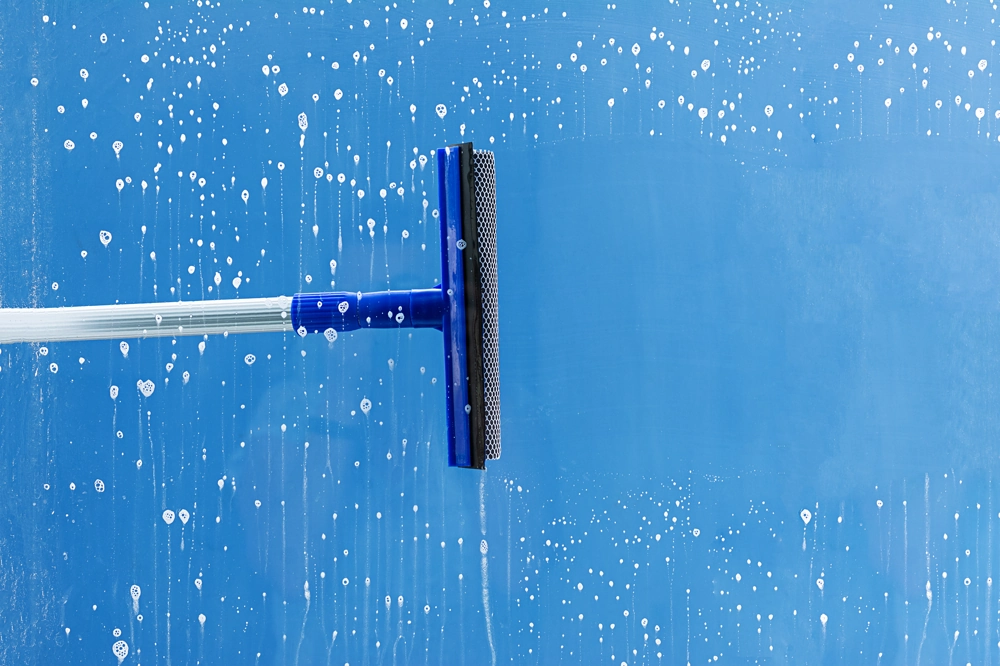 Fensterreinigung mit blauem Gummiwischer und Seifenlauge auf Glasoberfläche