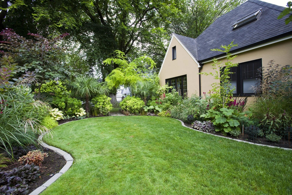Gartengestaltung mit geschwungenem Rasen, vielfältigen Staudenbeeten entlang der Hausfassade und gepflegter Grünfläche