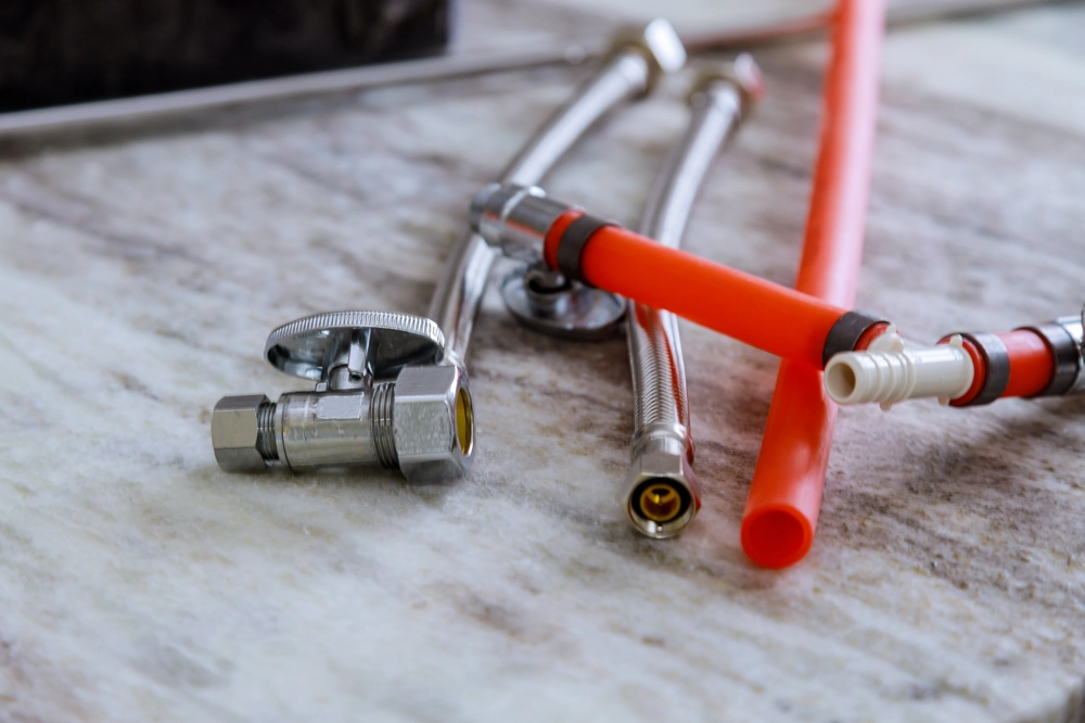Metallwinkelventil mit flexiblen Anschlussschläuchen und roten PEX-Rohren auf Marmorplatte – Sanitärinstallation