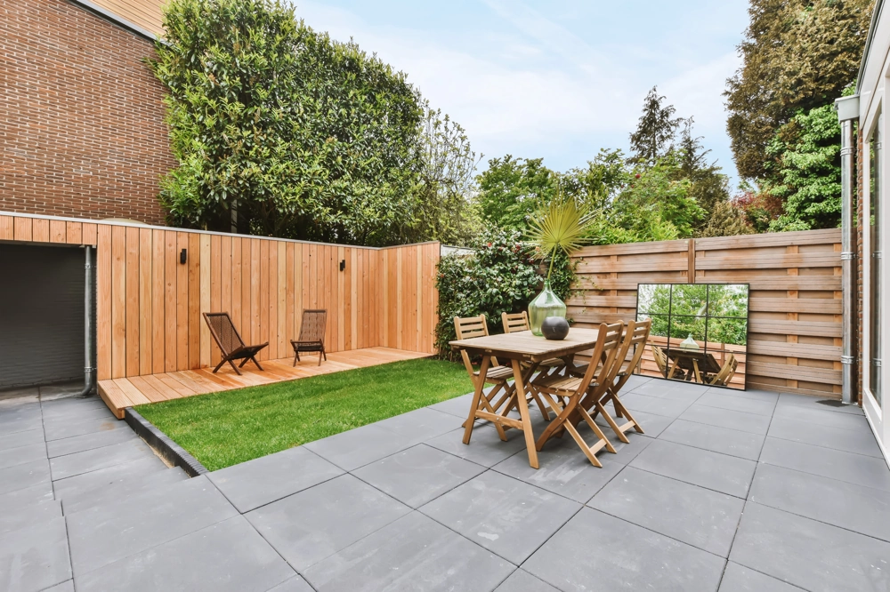 Terrassengestaltung mit großformatigen grauen Fliesen, Holzzaun, Gartentisch mit Stühlen und Rasenfläche